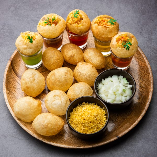 A wooden tray holds multiple pani puri, some filled with colorful chutneys, alongside bowls of toppings.