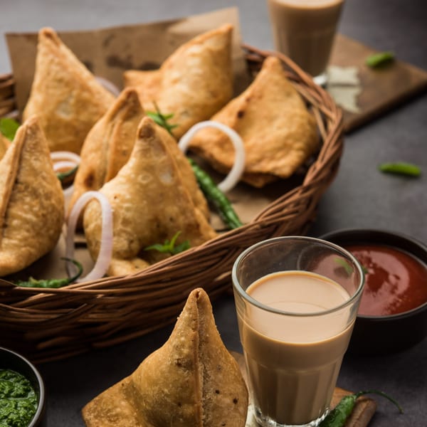 Golden-brown samosas in a woven basket with onion rings, green chilies, and two glasses of chai.